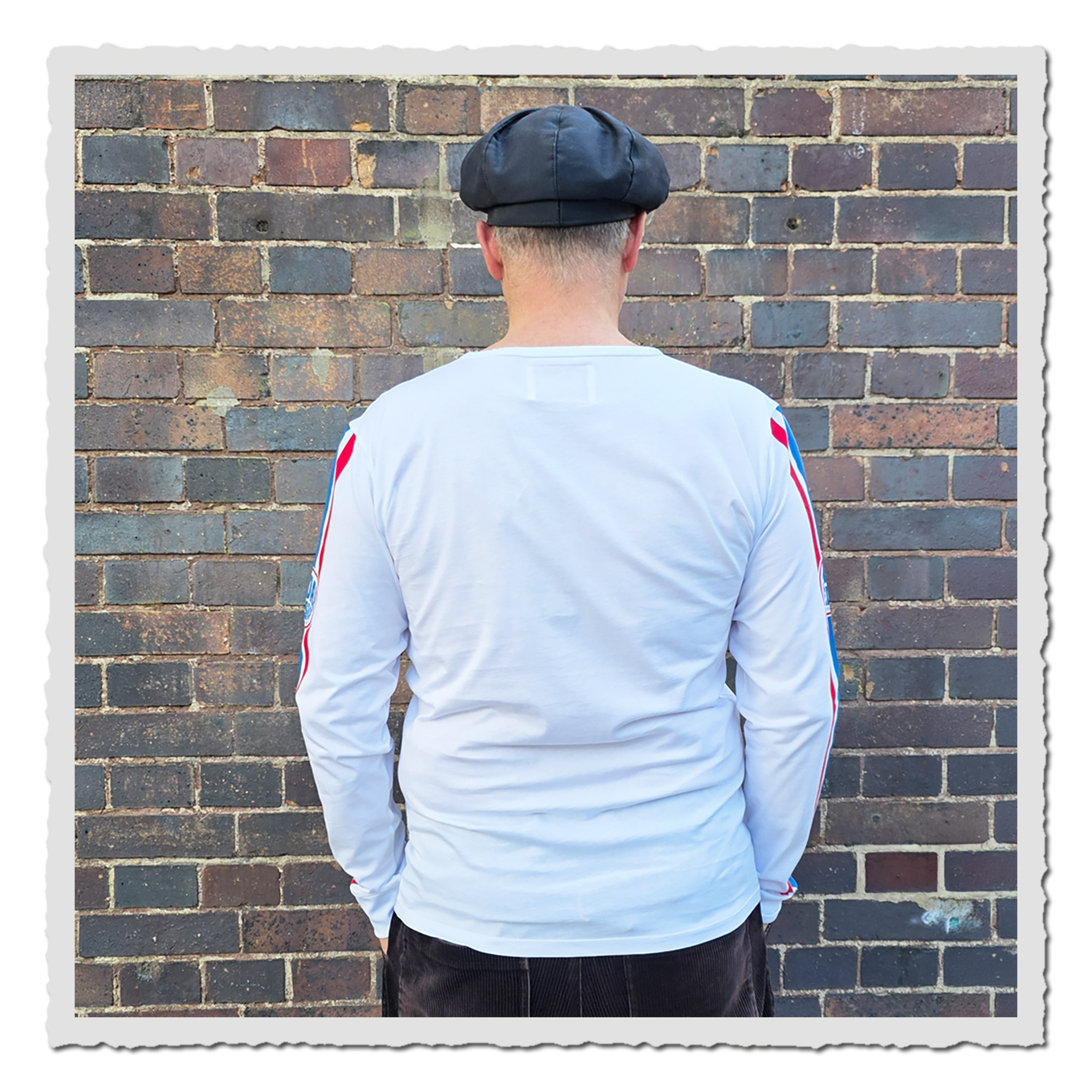 Rückansicht einer Person mit weißem Langarmshirt und rot-blauen Ärmelstreifen vor Backsteinwand.